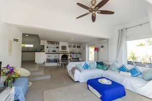 A spacious living area with a sofa set, a coffee table, and a dining table. There is a ceiling fan and a fireplace in the background.