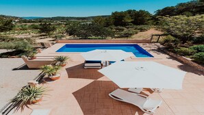 Ein rechteckiger Pool mit glasklar blauem Wasser, umgeben von einer gepflasterten Terrasse mit weißen Liegestühlen und Sonnenschirmen.