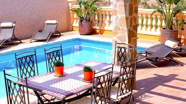 Un espace aménagé au bord de la piscine, avec une terrasse carrelée, des chaises longues, une table recouverte d’une nappe à motifs et des plantes en pot.