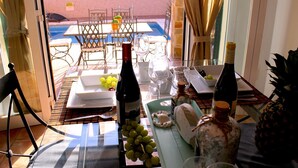 Un espace de restauration en plein air avec une table dressée pour deux personnes, agrémentée d’une bouteille de vin, de verres, d’un ananas et d’un bol de raisins.