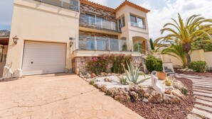 A two-story house with a garage, a balcony, and a landscaped garden with rocks and plants. - Modern Villa near Beaches (Roses)