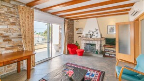 A spacious living room with a stone wall, wooden ceiling beams, a fireplace, and a large window. - Modern Villa near Beaches (Roses)