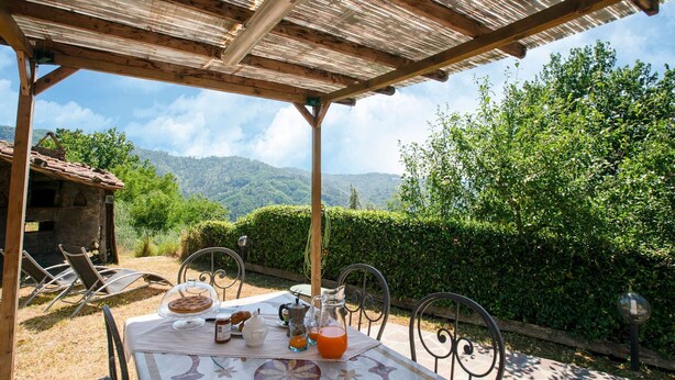 Ein Essbereich im Freien mit einem für das Frühstück gedeckten Tisch, umgeben von Stühlen, unter einer Holzpergola, mit Blick auf üppiges Grün und Berge im Hintergrund.