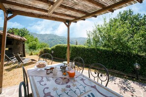 An outdoor dining area with a table set for breakfast, surrounded by chairs, under a wooden pergola, with a view of lush greenery and mountains in the background. - Belvilla by OYO Casa di Laino (Lucca)