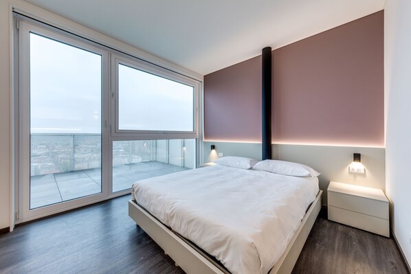 A modern bedroom with a large window, a bed, a nightstand, and a wall-mounted light.