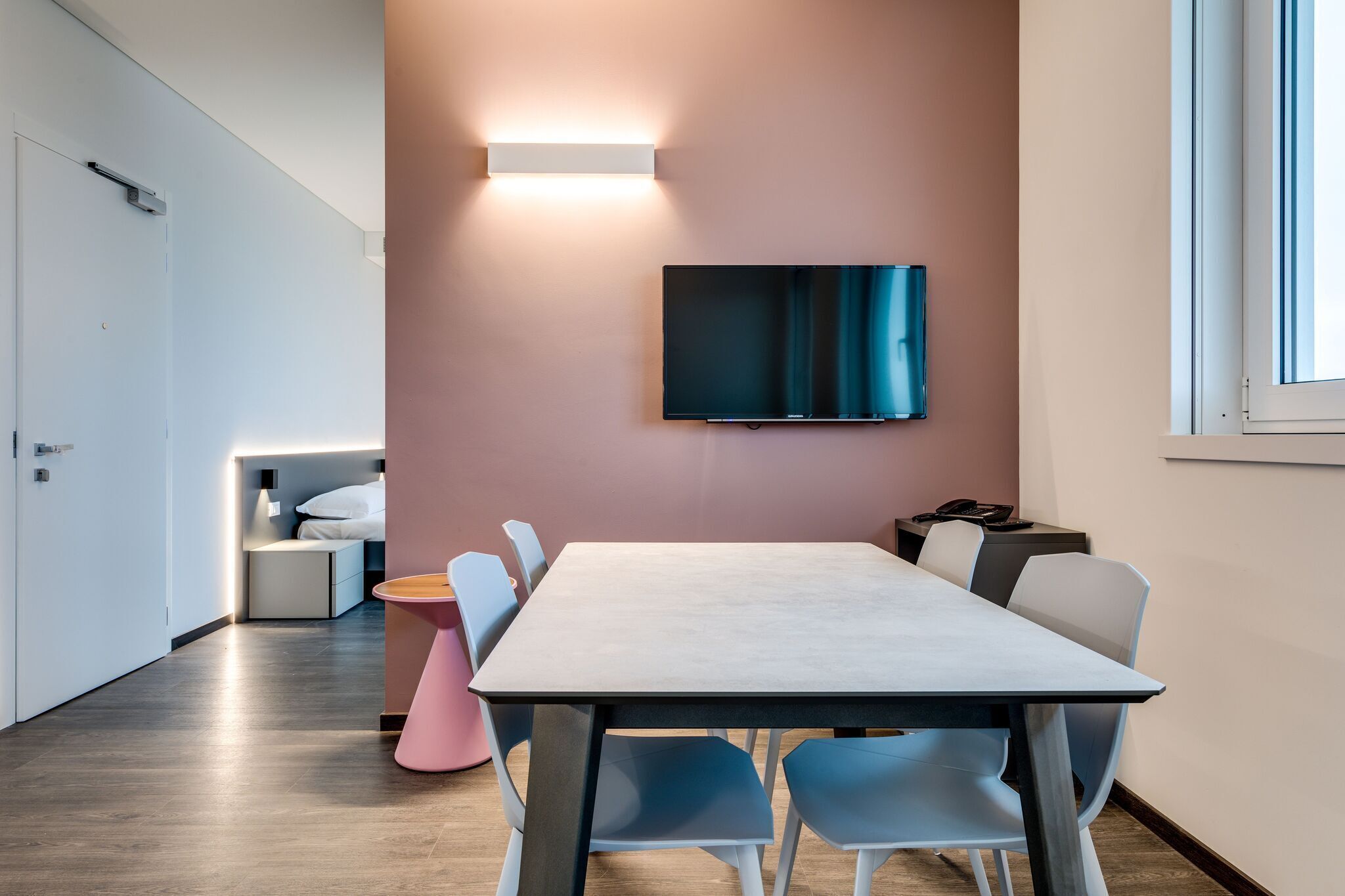 Una sala de reuniones moderna con una larga mesa, sillas azules, una mesita auxiliar rosa y una televisión montada en la pared.