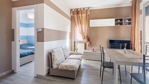 A modern, well-lit room with a bed, sofa, dining table, and a small kitchenette area.