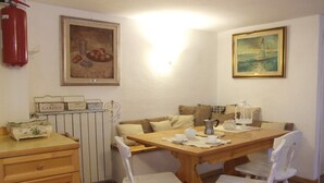 Uma área de refeições com uma mesa de madeira, cadeiras brancas, um radiador e duas pinturas nas paredes.