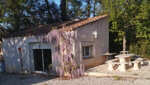 Un edificio pequeño con un camino de grava, un patio con una mesa y bancos, y una flor púrpura colgante.