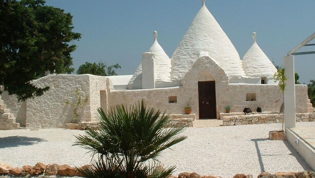 A white building with a conical roof and a central door, surrounded by gravel and a palm tree.