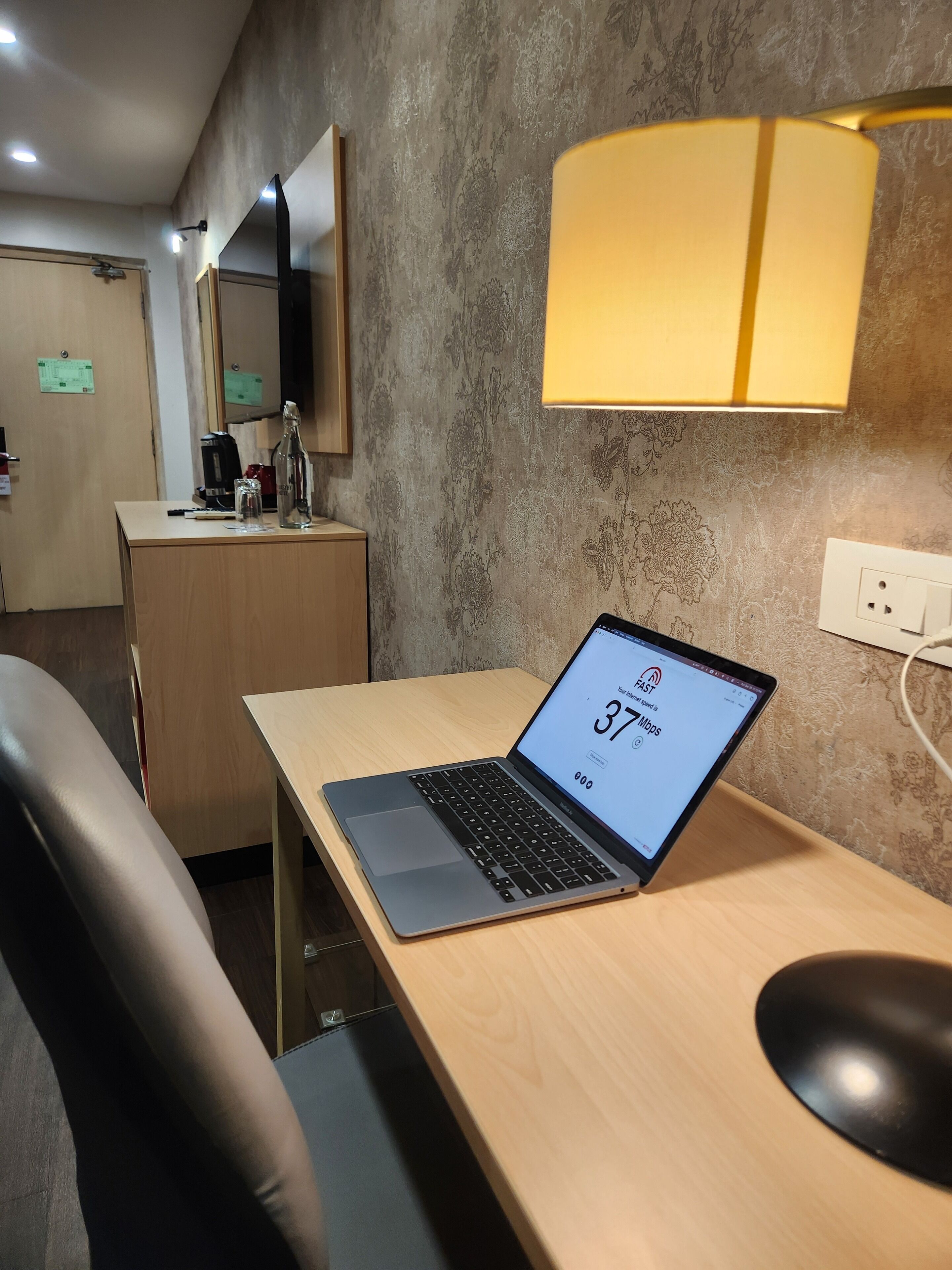 Una habitación de hotel moderna con un escritorio de madera, una computadora portátil mostrando los resultados de una prueba de velocidad, una lámpara y un espejo.
