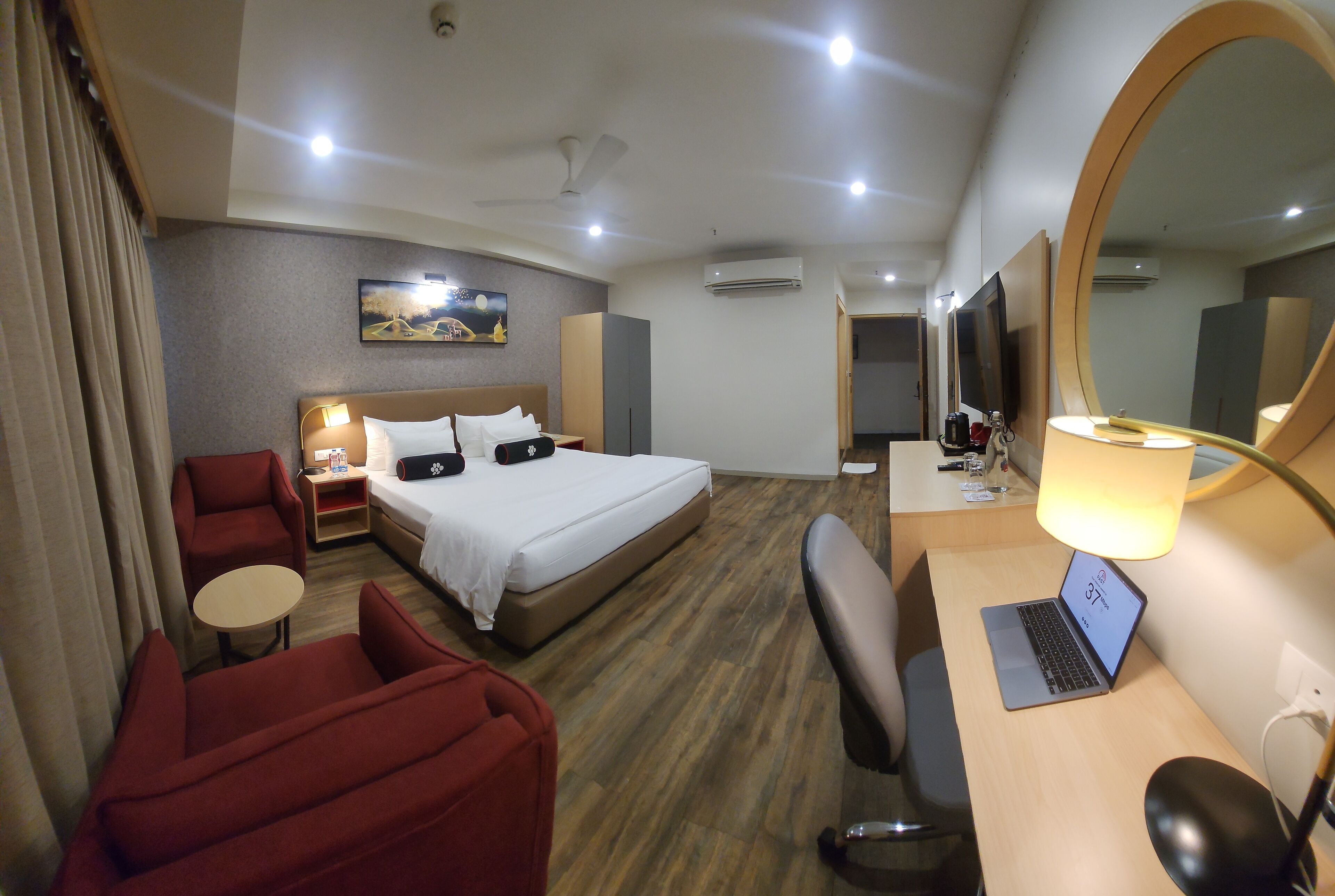 Una habitación de hotel con una cama, un escritorio con una computadora portátil, una silla, una mesita, un sofá, un televisor y un espejo grande.