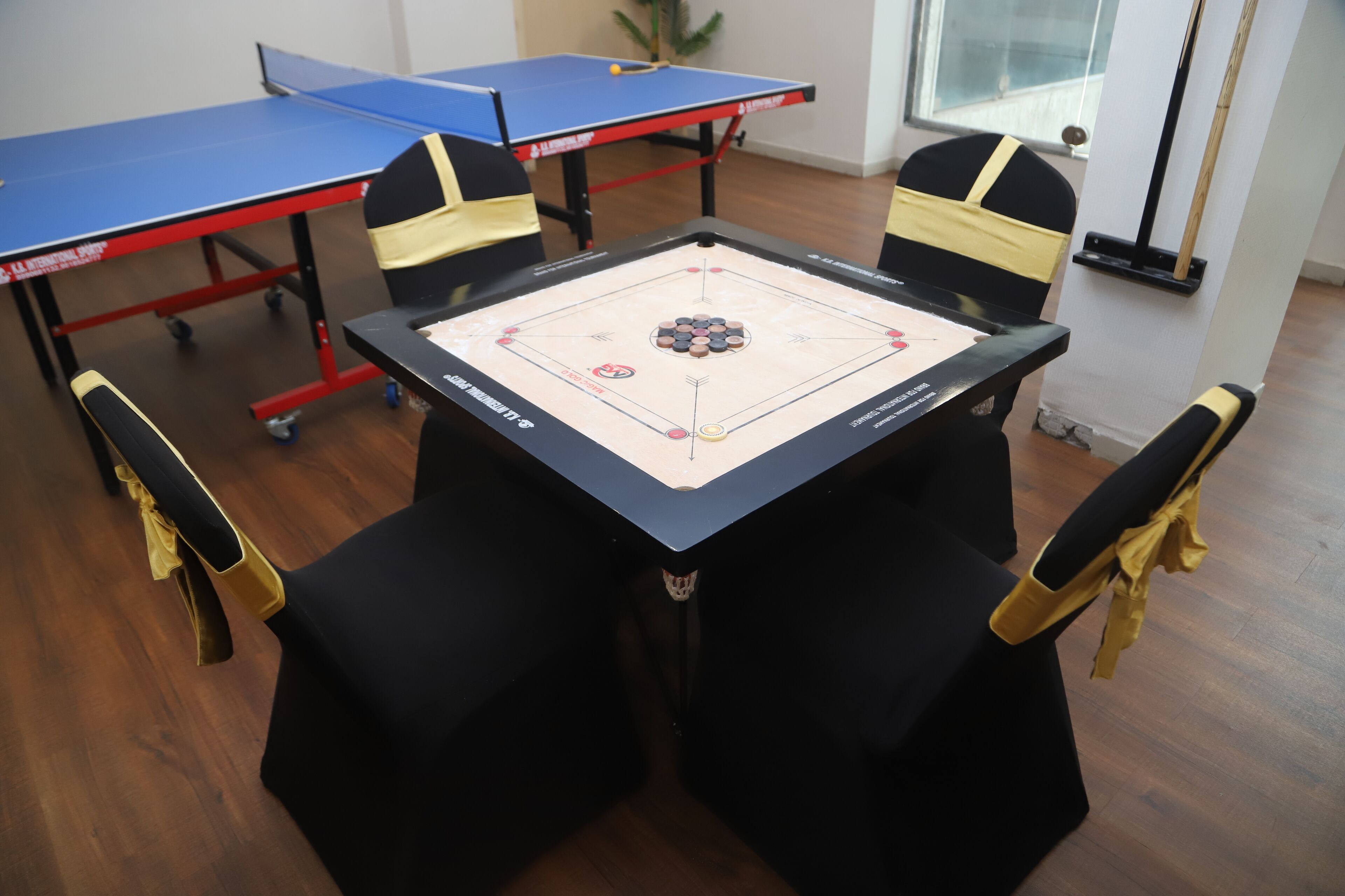 A table with a board game, four chairs, and a blue table tennis table in a room with wooden flooring.