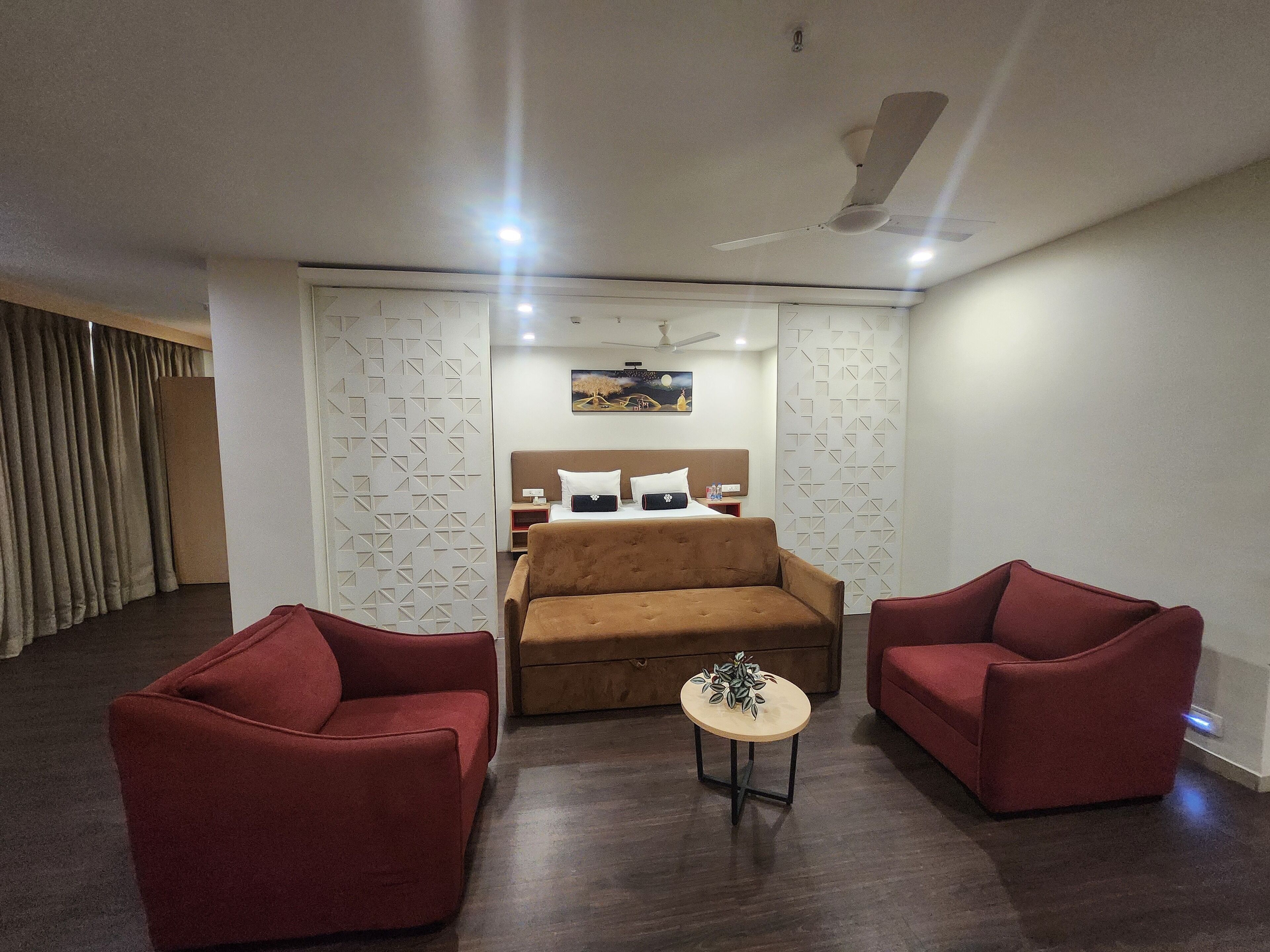 Une chambre d’hôtel moderne avec un lit, deux fauteuils rouges, une petite table ronde et un ventilateur de plafond.