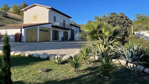 Una casa de dos pisos con balcón, rodeada por un camino de grava y un jardín con diversas plantas.