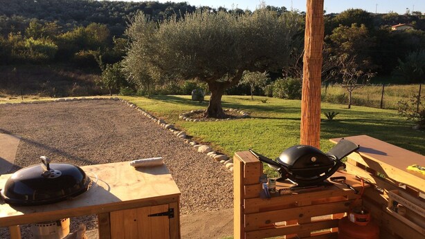 Una parrilla de barbacoa montada sobre una mesa de madera en un área exterior con césped, con un árbol y una colina al fondo.