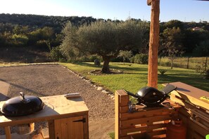 Una parrilla de barbacoa montada sobre una mesa de madera en un área exterior con césped, con un árbol y una colina al fondo.