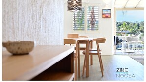 A modern dining area with a wooden table and chairs, a hanging light fixture, and a view of the outdoors through a window with blinds. - St Tropez 10 - Classic Coastal Water Views (Noosa Heads)