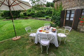 A round table with white chairs set on a lawn, surrounded by a garden with a stone wall and greenery.