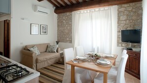 Una zona de estar acogedora con una pared de piedra, techo de madera, una mesa de comedor para dos, un sofá con cojines y un televisor de pantalla plana sobre un mueble.
