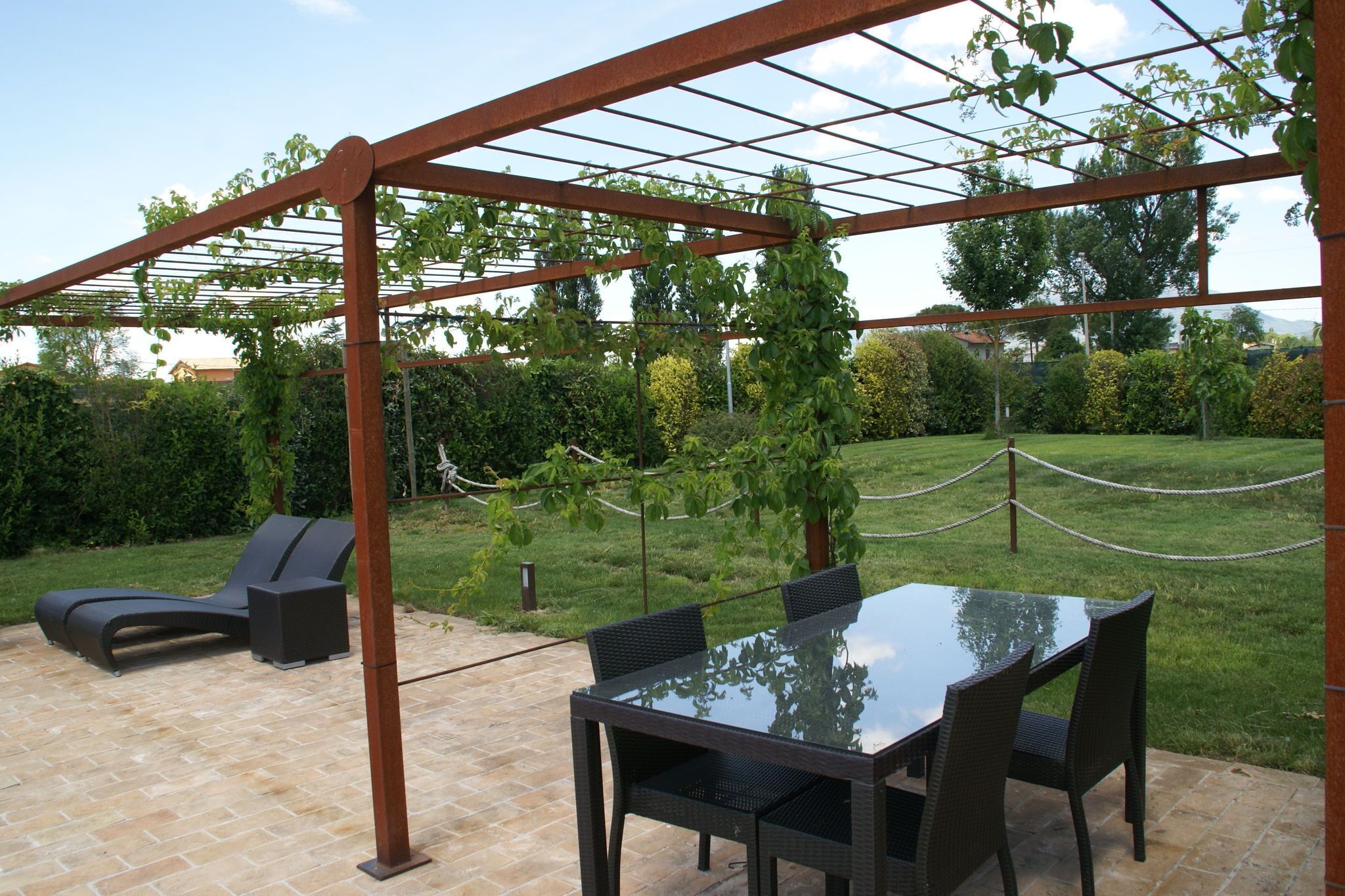 An outdoor patio with a glass table and chairs, a lounge chair, and a pergola with climbing plants.