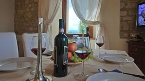 Una mesa de comedor dispuesta con copas de vino, una botella de vino, una botella de agua, un cuenco de fruta y un televisor montado en la pared.