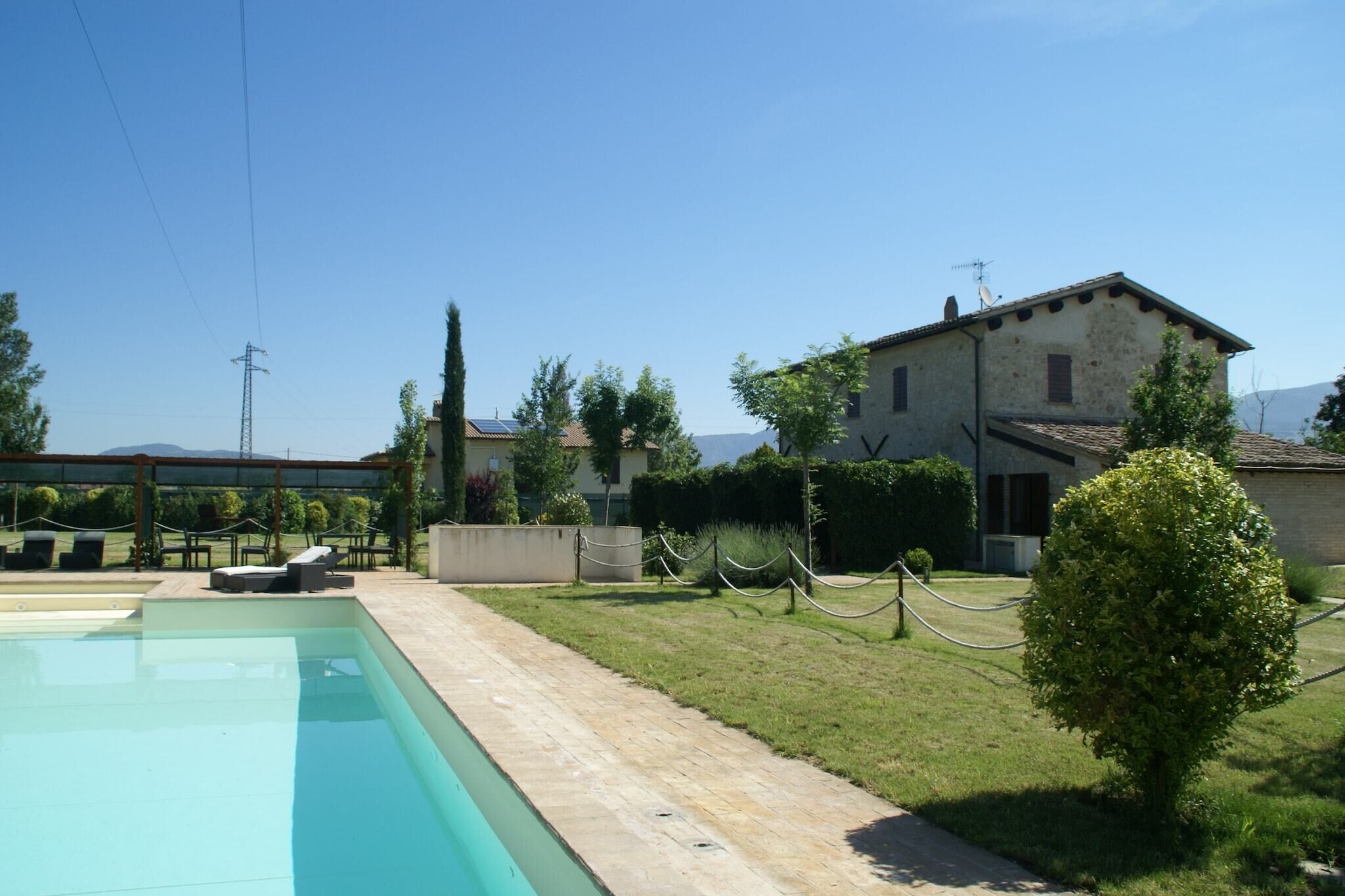 Un espace piscine avec un coin salon, un bâtiment doté d’un toit en tuiles et une pelouse verdoyante bordée d’une haie.