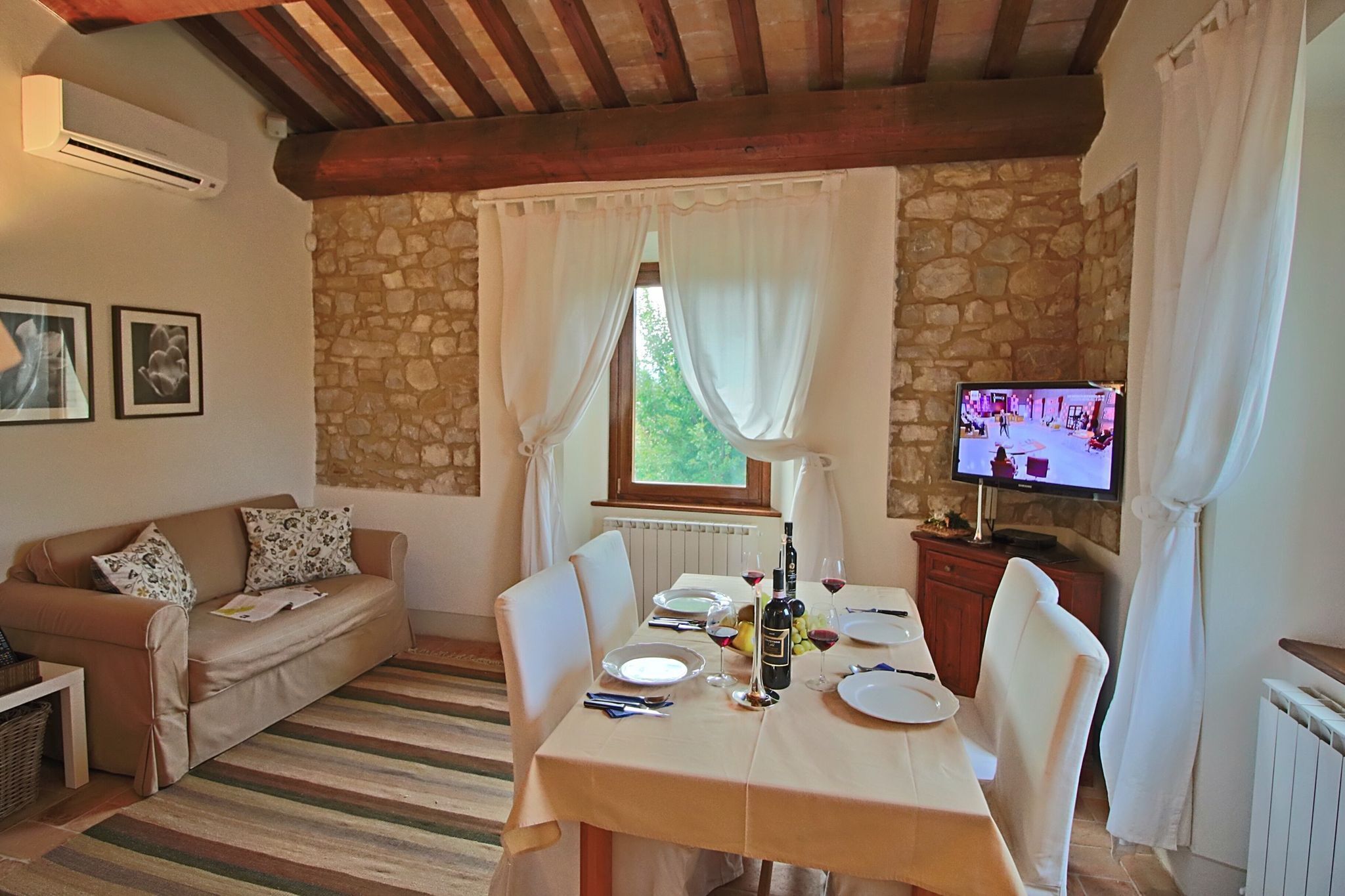 Un coin repas chaleureux avec un mur en pierre, un plafond en bois, une table dressée pour deux personnes et un téléviseur posé sur un meuble.