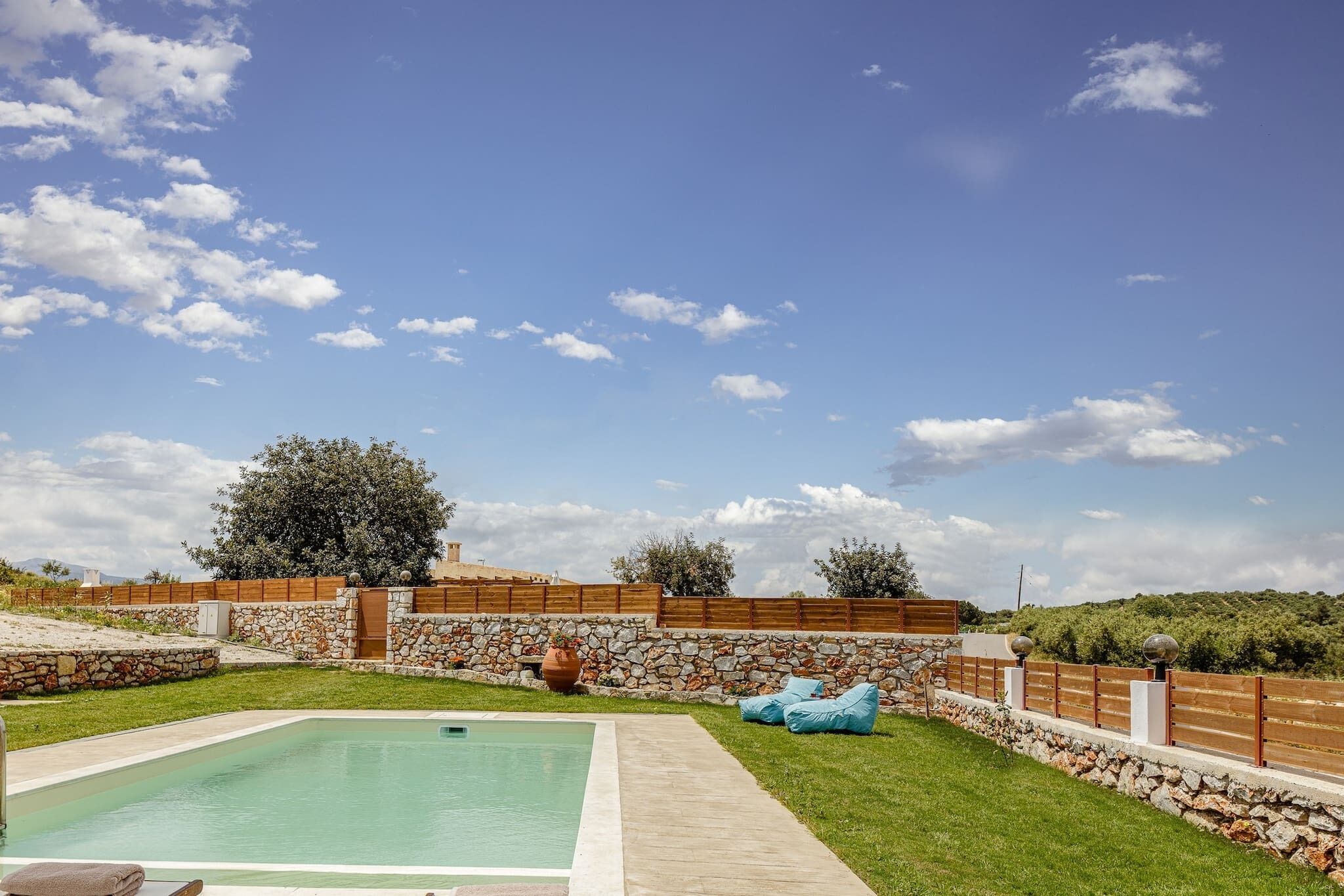 A swimming pool with a stone wall and wooden fence, surrounded by grass and trees.