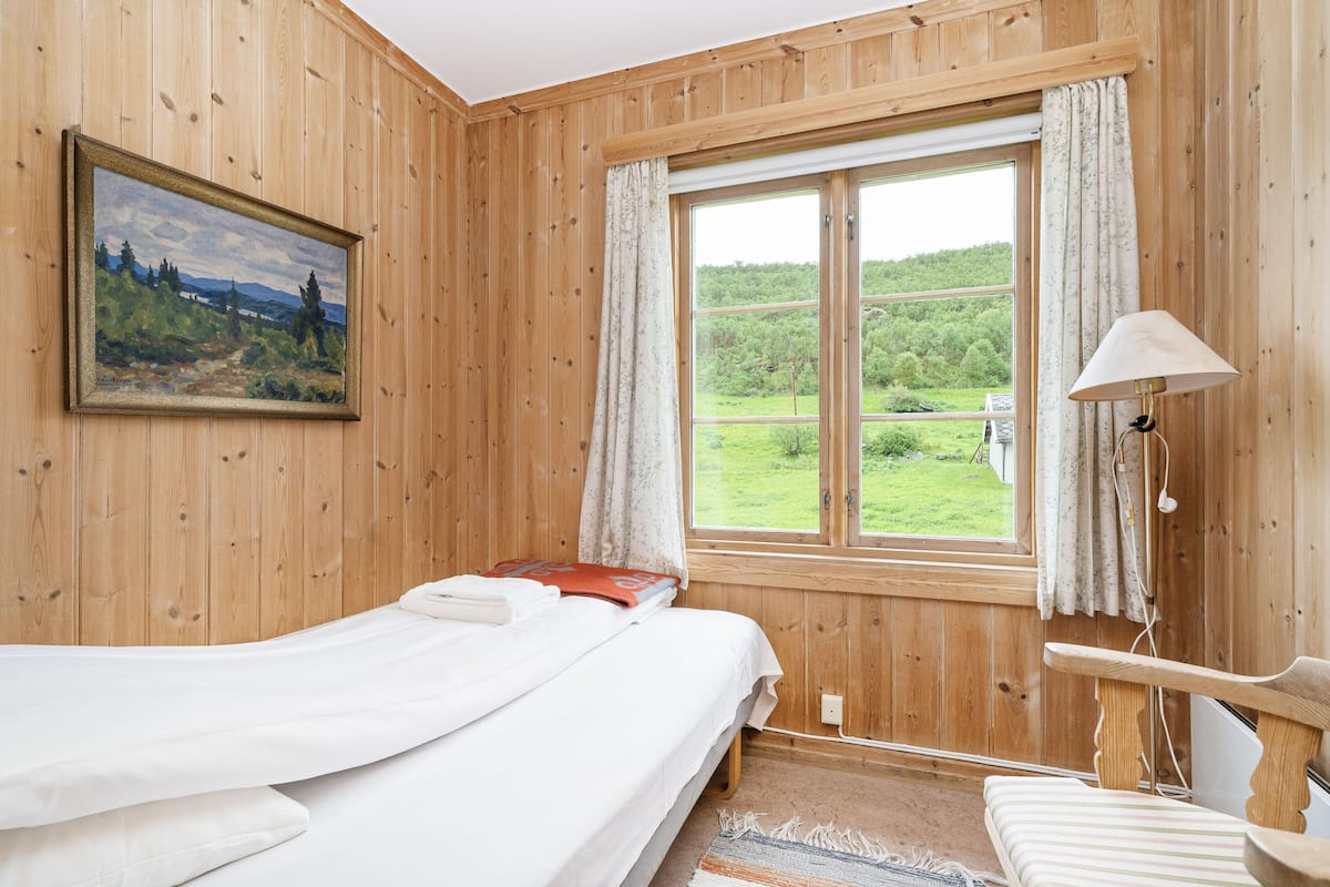 Una habitació de fusta amb un llit, un quadre, una finestra amb cortines i una làmpada.