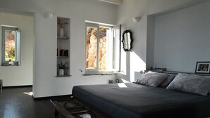 Une chambre avec un grand lit, un banc en bois, une fenêtre donnant sur des falaises et une étagère avec des livres et un vase.