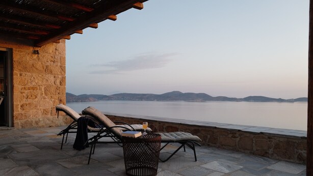 Une terrasse en pierre avec des chaises longues et une petite table, offrant une vue sur une étendue d’eau calme et des collines lointaines.
