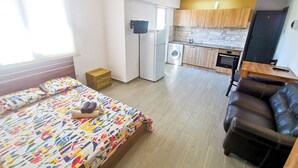 A compact room with a bed, a small wooden nightstand, a black leather chair, a washing machine, and a kitchenette with wooden cabinets and a tiled backsplash. - Hidden Gem City Studio (Larnaca)