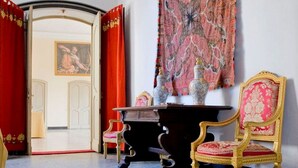 A room with a large red curtain, a patterned tapestry, a dark wooden table, and two ornate chairs.