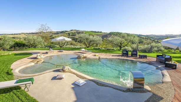 A swimming pool with a curved design, surrounded by a deck with outdoor furniture and a view of a hilly landscape.
