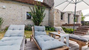 A wooden deck with two lounge chairs and a table, a stone building with a window, and a large umbrella. - Farmhouse in Yèvre-le-Châtel with Heated Pool (Yèvre-la-Ville)
