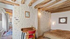 A bedroom with a stone wall, wooden beams, a desk with a red chair, a bed with a patterned blanket, and a small mirror on the wall. - Farmhouse in Yèvre-le-Châtel with Heated Pool (Yèvre-la-Ville)