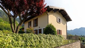 A two-story house with a stone wall and a metal fence, surrounded by lush greenery and mountains in the background. - Holiday Home in Idro near Scenic Hills (Idro (bs))