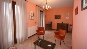 A room with two orange chairs, a glass table, a chandelier, a radiator, a television, and a wooden cabinet. - Holiday Home in Idro near Scenic Hills (Idro (bs))