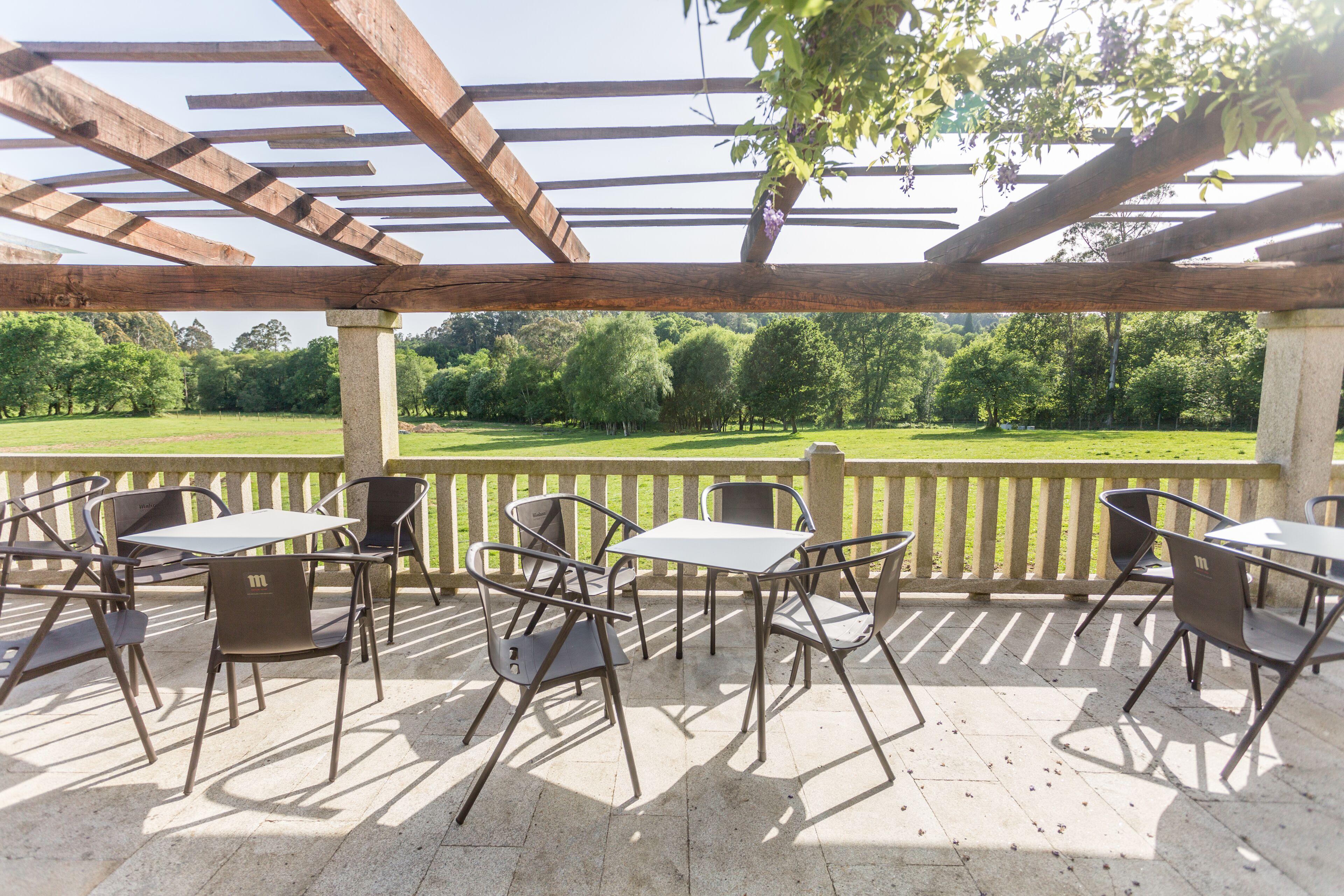 An outdoor patio with metal tables and chairs, a wooden pergola, and a view of a green field.