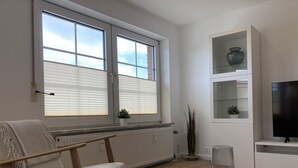 A modern living room with a white sofa, a coffee table, a TV on a stand, and a window with blinds. - Apartment house "Charibi" a well-kept residential complex near the beach (Dahme)