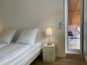 Une chambre avec un lit, une table de chevet avec une lampe, et une vue sur une terrasse extérieure avec des chaises et un mur en briques.