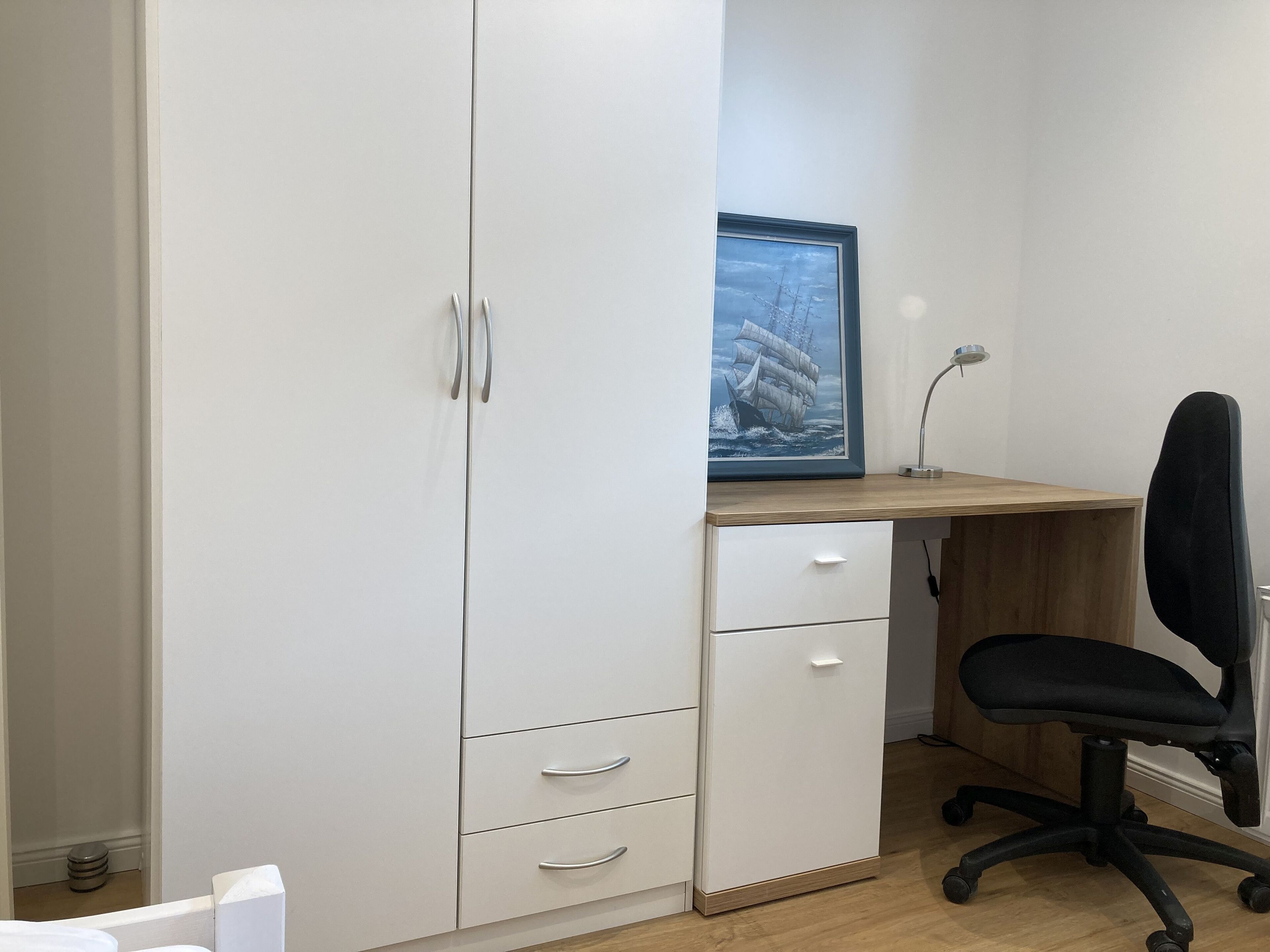 Een kamer met een witte kast, een houten bureau, een zwarte bureaustoel en een lijst met een afbeelding van een schip aan de muur.
