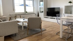 A modern living room with a white sofa, a dining table, and a TV. - Apartment house "Charibi" a well-kept residential complex near the beach (Dahme)