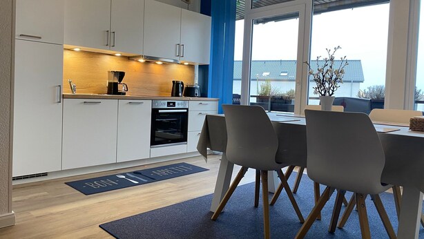 A modern kitchen with white cabinets, a built-in oven, and a coffee machine. There is a dining area with a table and chairs, and a view of the outside through large windows. - Maisonette apartment near the beach - beach chair included! (Dahme)