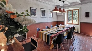Un espace repas avec une longue table dressée pour plusieurs personnes, des chaises, des luminaires suspendus, des œuvres d’art encadrées accrochées aux murs et une fenêtre donnant sur un paysage verdoyant.
