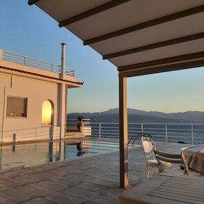 Un espace aménagé au bord de la piscine, offrant une vue sur la mer et les montagnes, un coin salon couvert et un bâtiment doté d’une cheminée.