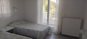 A small, simple room with two beds, a radiator, and a window with a view of the outdoors. - Charming 4-bed Villa in Pefkali (Corinth)
