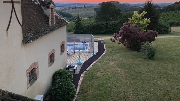 A house with a sloped roof, a small garden with a hedge and flowers, and a view of the sunset over rolling hills. - Country house with swimming pool (Coulx)