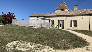 A two-story building with a tiled roof, a covered patio area with chairs, and a gravel path leading to the entrance. - Country house with swimming pool (Coulx)
